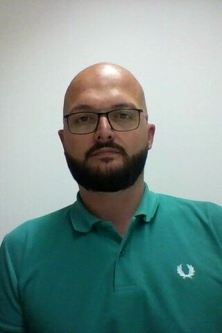 Portrait photo of a bald man with glasses and a beard wearing a green polo shirt, facing forward with a neutral expression