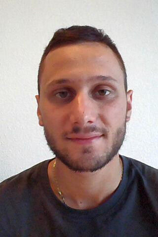 Portrait photo of a young man with short dark hair and a trimmed beard wearing a dark t-shirt, facing forward with a neutral expression